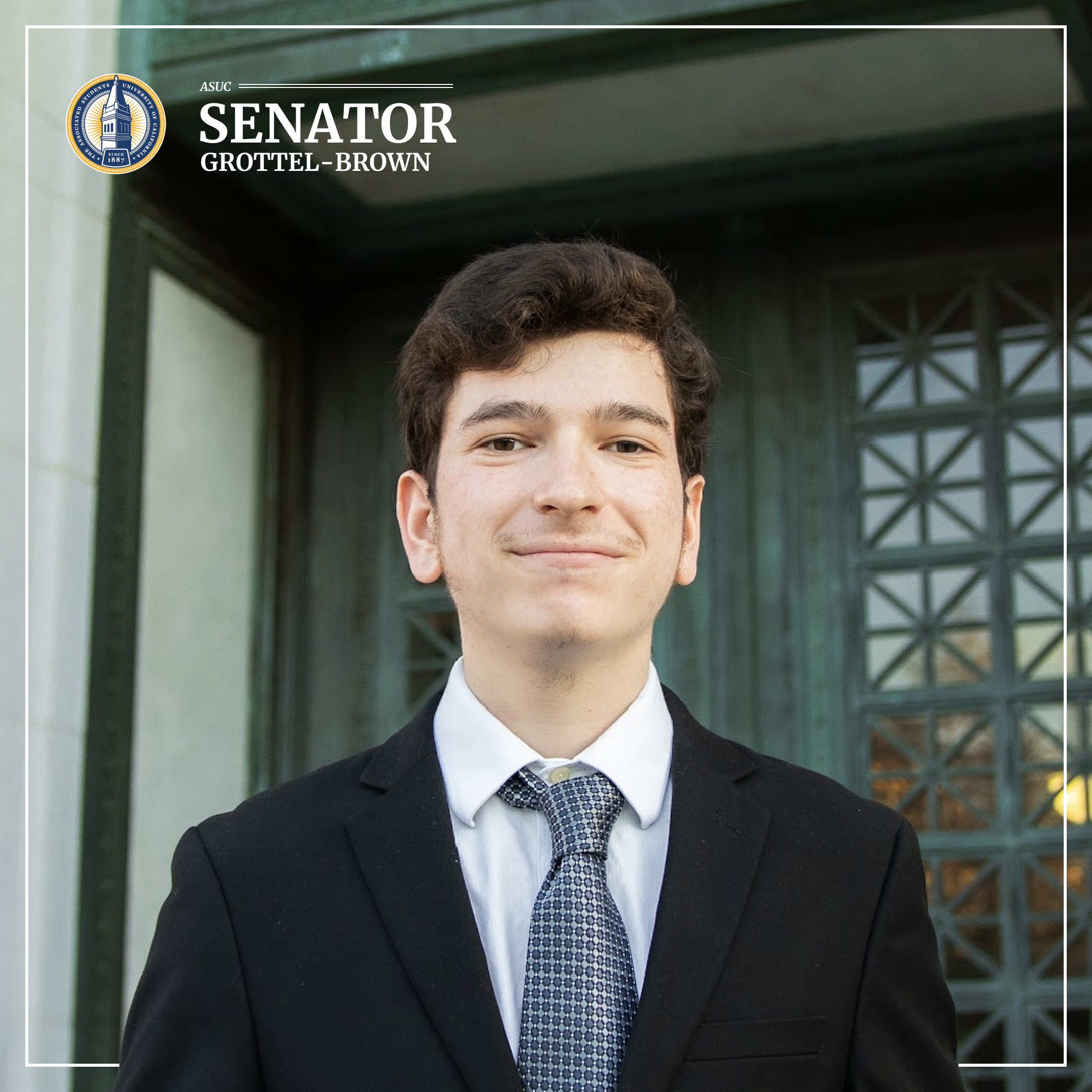 Gabe looks dashing wearing a black suit and red tie. He's standing in front of bright green foliage, and we can make out the faintest outline of a modern building in the background.