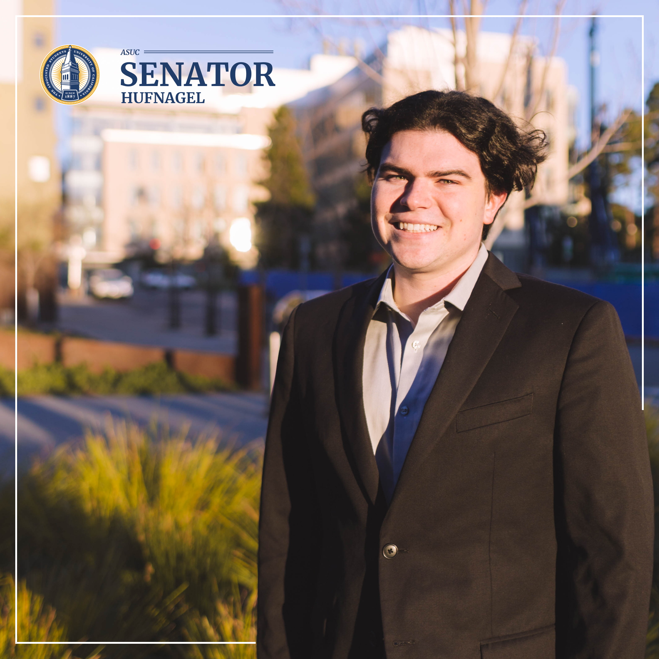 Gabe looks dashing wearing a black suit and red tie. He's standing in front of bright green foliage, and we can make out the faintest outline of a modern building in the background.